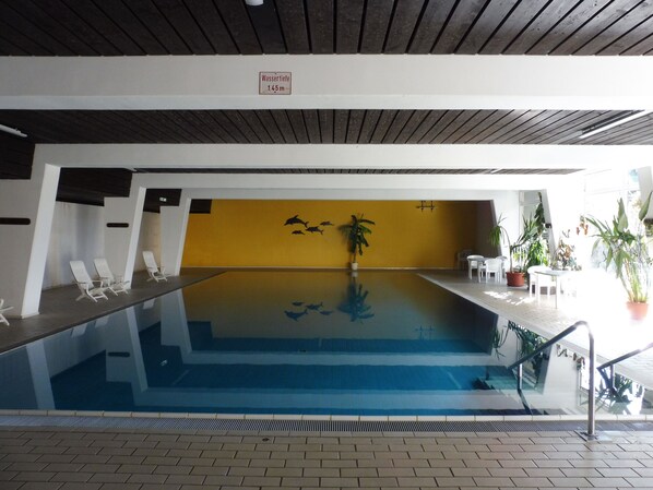 An indoor swimming pool with a yellow wall, lounge chairs, and potted plants.