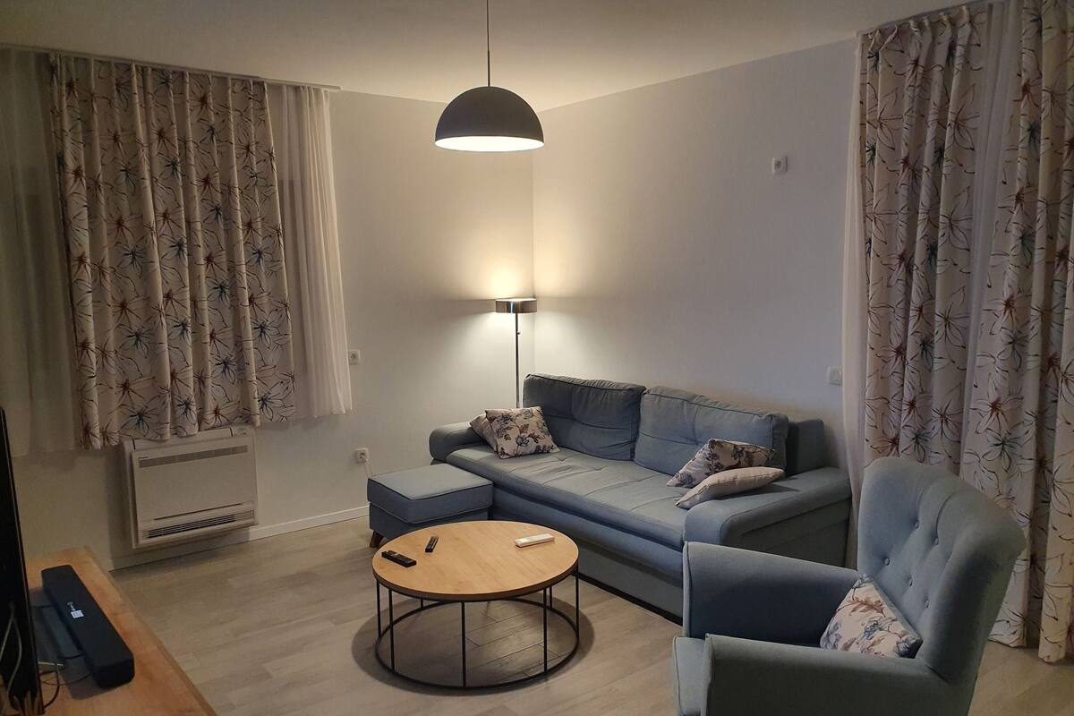 A modern living room with a grey sofa, a round coffee table, and a blue armchair. There is a floor lamp, a radiator, and curtains with a floral pattern.