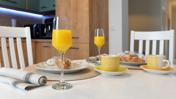 A breakfast setting with two glasses of orange juice, plates with croissants, and a newspaper on a table. - Q apartments (Murter-Kornati)