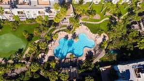 Eine Luftaufnahme eines Resorts mit einem einzigartigen, wellenförmigen Pool, umgeben von Palmen und einem Golfplatz.