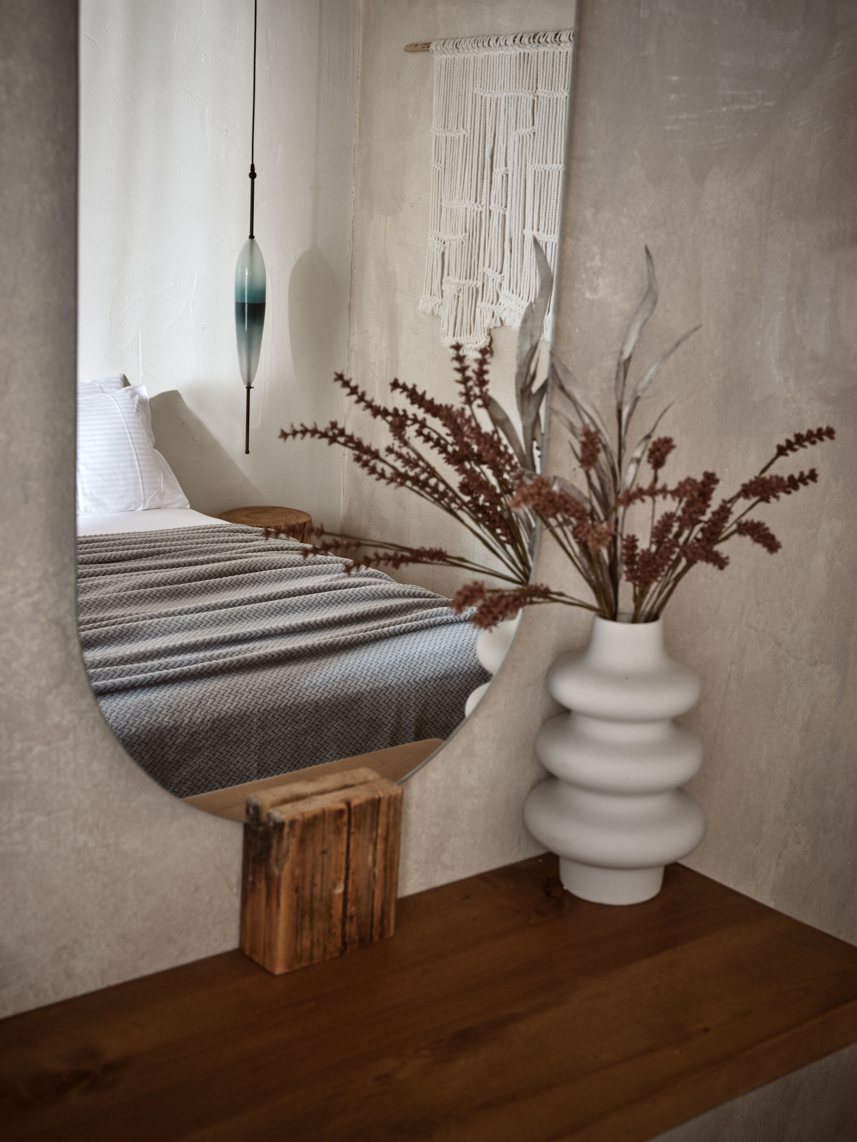 A bedroom with a bed, a white vase with dried plants, a wooden stool, and a macrame wall hanging.