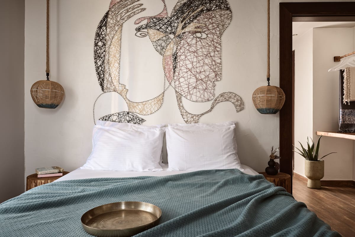 A bedroom with a large abstract wall art, a bed with white bedding, a blue blanket, and a wooden floor.