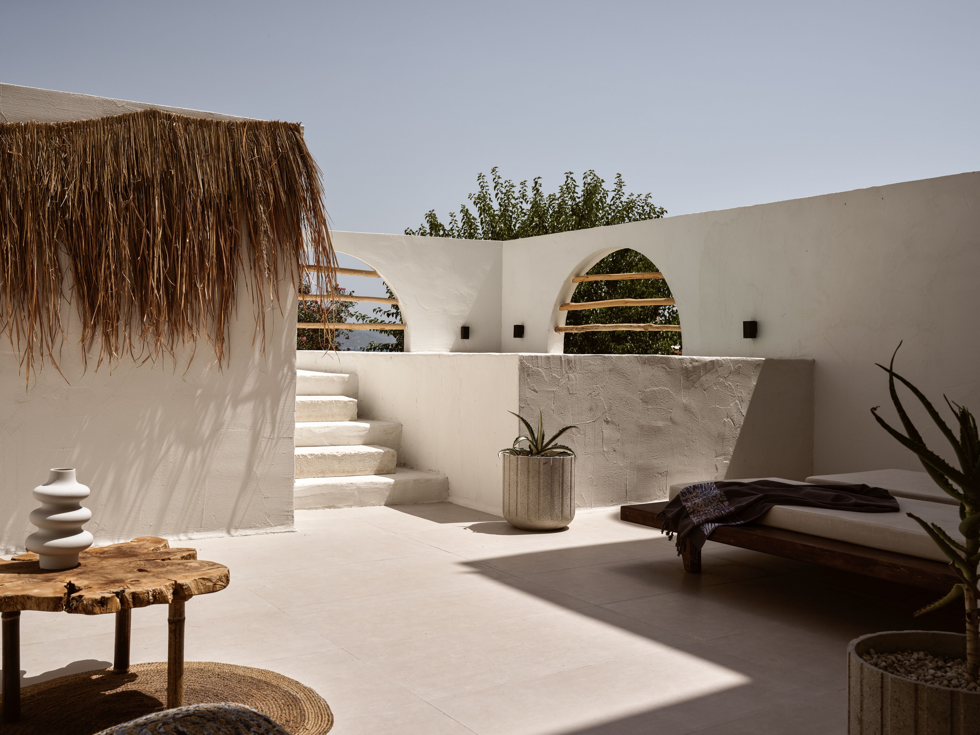 A sunlit outdoor patio with a thatched roof, a staircase, a seating area, and potted plants.