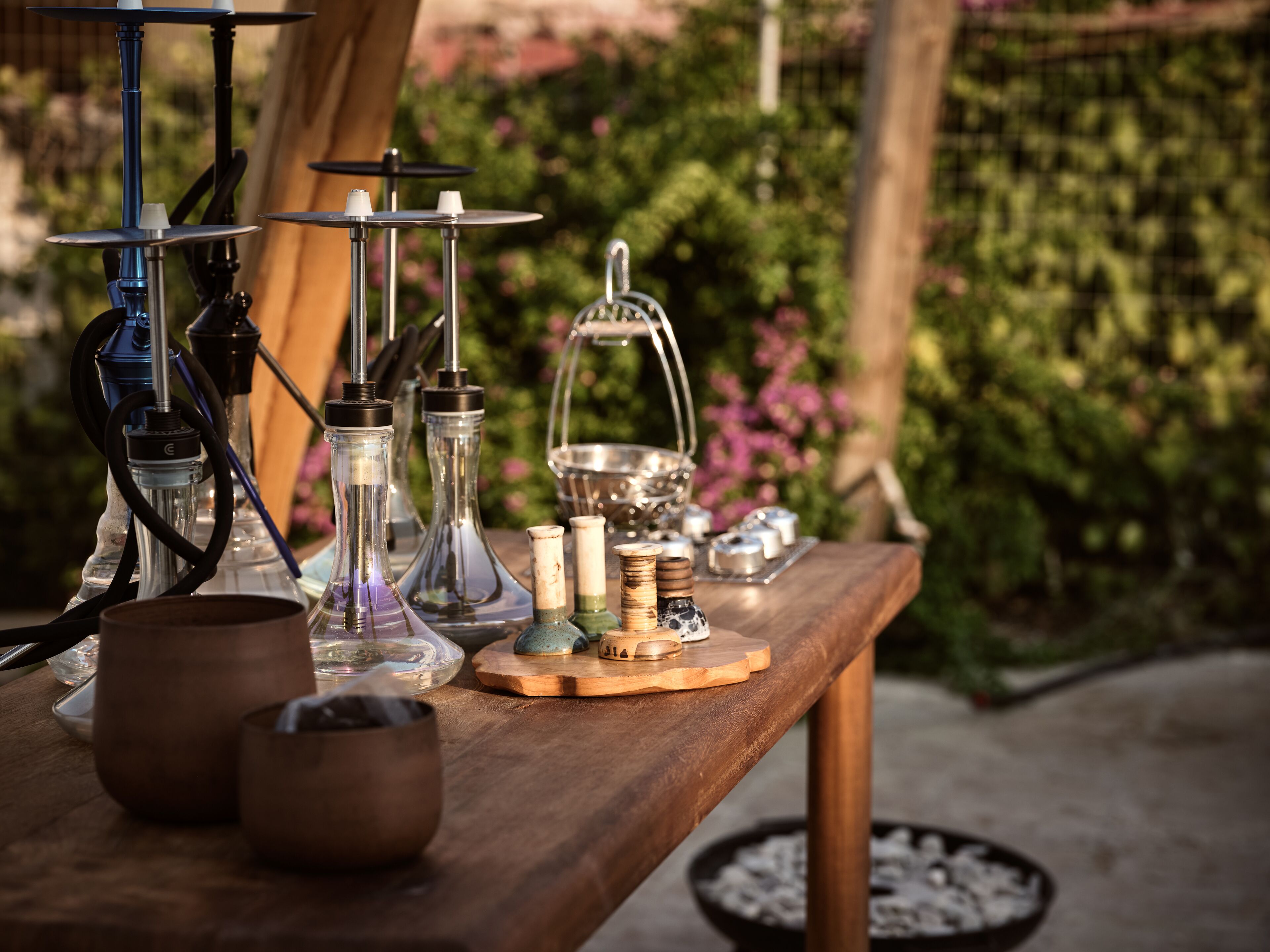 A wooden table with various smoking accessories, including hookahs and pipes, set outdoors with greenery in the background.