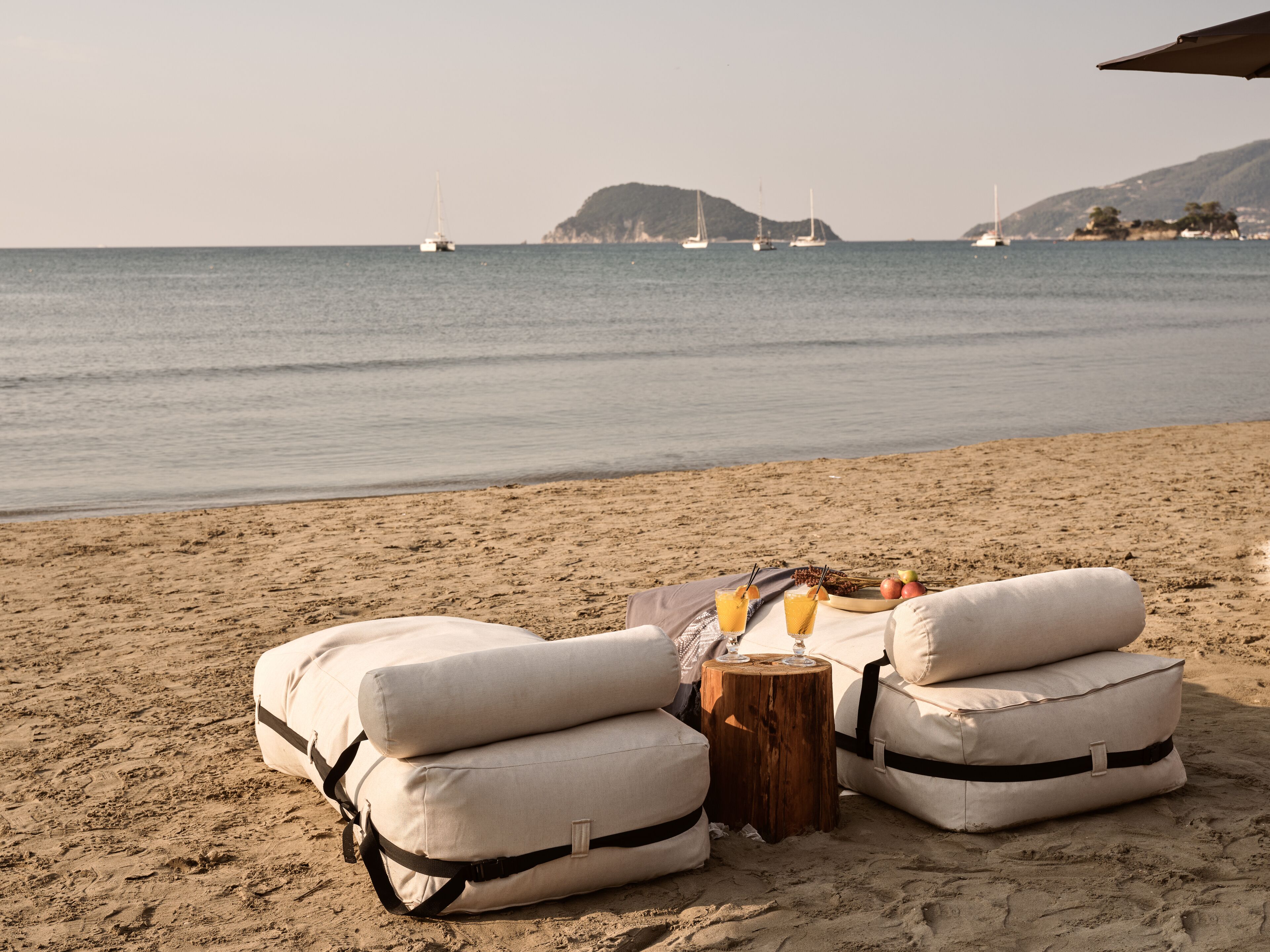 Een strandsetting met twee beige lounge stoelen, een klein houten tafeltje met drankjes en een uitzicht op de zee met boten in de verte.