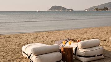 Een strandsetting met twee beige lounge stoelen, een klein houten tafeltje met drankjes en een uitzicht op de zee met boten in de verte.