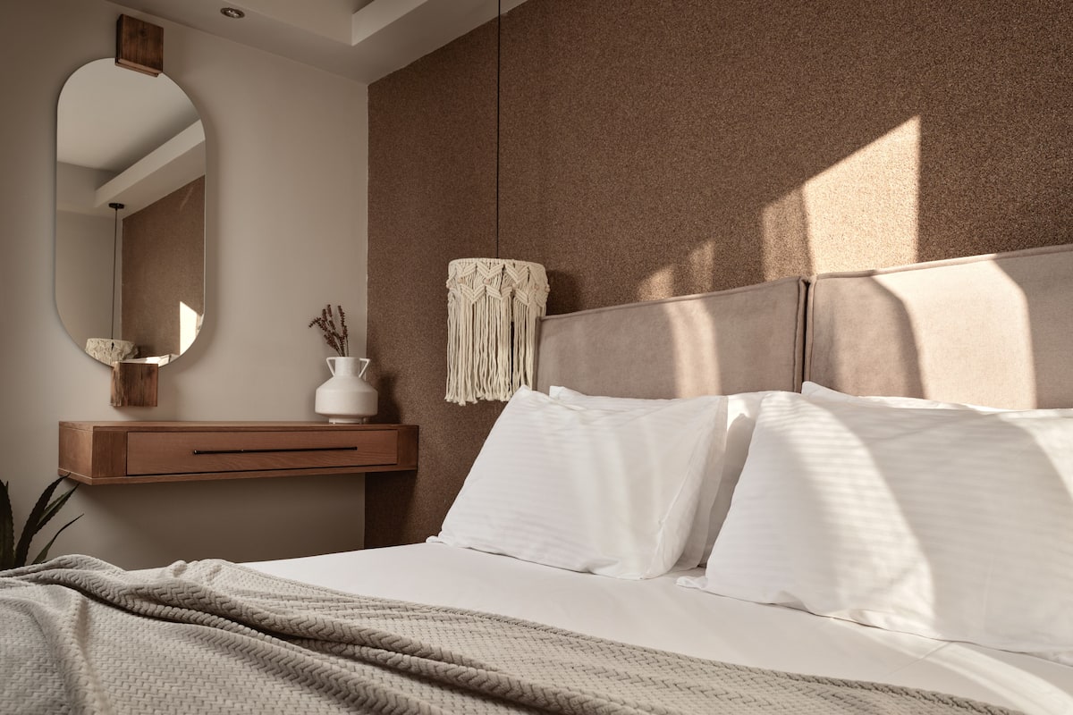 A bedroom with a bed, a bedside table, a mirror, and a macrame wall hanging.
