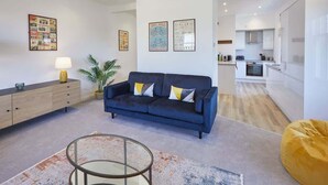Ein modernes Wohnzimmer mit einem blauen Sofa, einem Glastisch und einem gelben Sitzsack. Es gibt eine Holzkommode mit einer Lampe, einer Topfpflanze und gerahmten Bildern an der Wand.