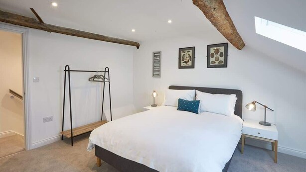 Ein Schlafzimmer mit einem großen Bett, einem Nachttisch mit Lampe, einer Holzbank, einem Kleiderständer und zwei gerahmten Bildern an der Wand.
