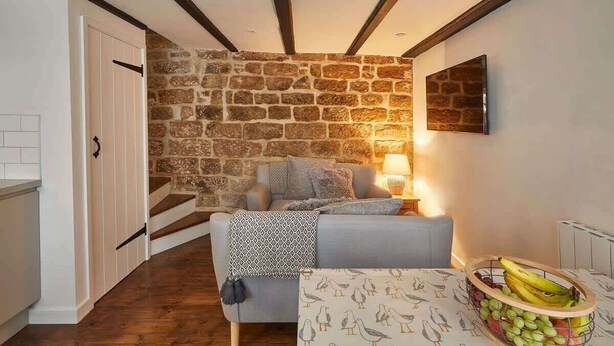 A cozy room with a stone wall, a bed with pillows, a table with a fruit bowl, and a staircase. - Host & Stay | Dolphin Cottage 1736 (Whitby)