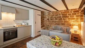 A compact kitchen with white cabinets, a built-in oven, and a stone wall. There's a dining area with a table covered by a bird-patterned tablecloth and a bowl of fruit. - Host & Stay | Dolphin Cottage 1736 (Whitby)