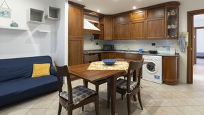 Cocina funcional con muebles de madera, una mesa de comedor con sillas, lavadora y un sofá azul con un cojín amarillo.