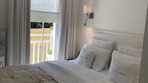 Una cama bien hecha con sábanas y almohadas blancas, una lámpara de noche y vistas a un balcón con cortinas.