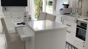 Una cocina moderna con una isla central, armarios blancos, un televisor de pantalla plana y un amplio ventanal con vistas a zonas verdes.