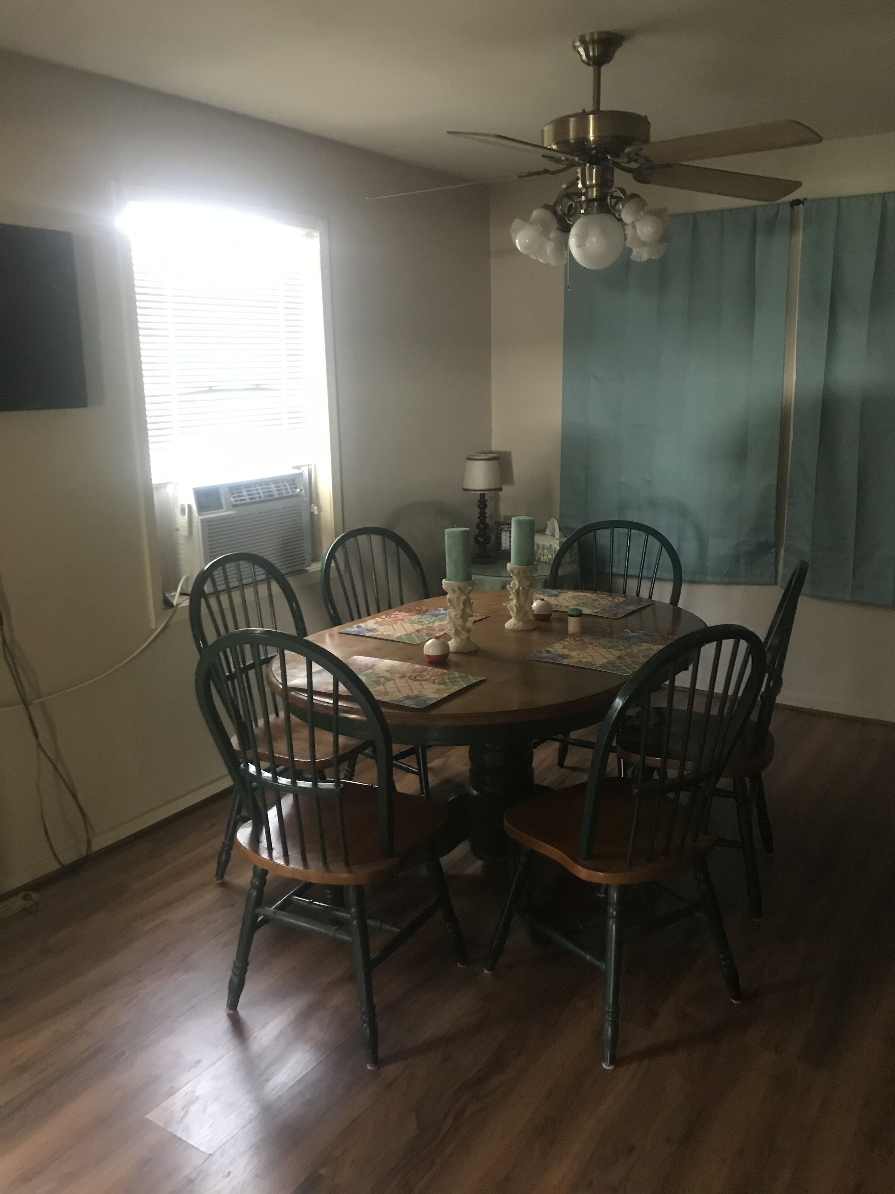 A dining room with a round wooden table, six chairs, a ceiling fan, and a window with blinds.