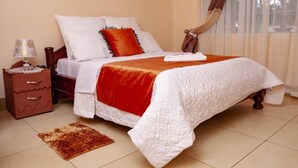 A neatly made bed with white and red bedding, a small bedside table with a lamp, and a brown rug on the floor. - Rest Guest House (Harare)