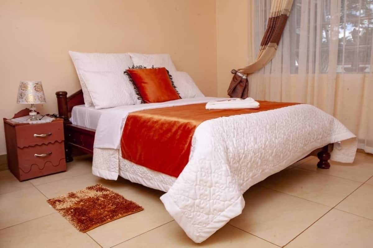 A neatly made bed with white and red bedding, a small bedside table with a lamp, and a brown rug on the floor.