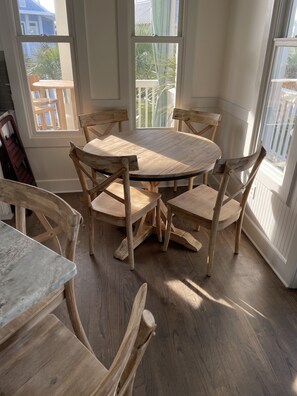 A dining area with a round wooden table and six chairs, large windows with white frames, and a view of a balcony with a railing.
