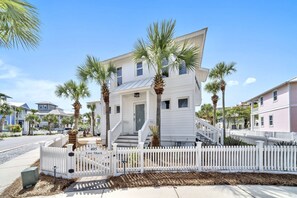 A white two-story house with a porch, palm trees, and a white picket fence.