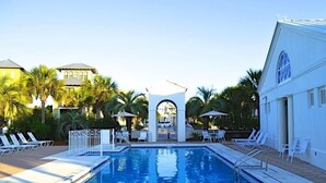 A swimming pool with lounge chairs and umbrellas, surrounded by palm trees and a building with a white facade.