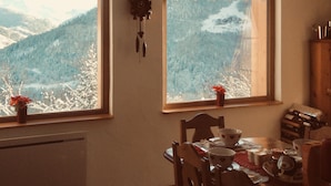 Un coin repas avec une table dressée pour deux personnes, une horloge au mur et une vue sur les montagnes enneigées par les fenêtres.