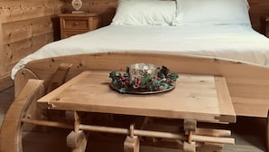 Une chambre dans une cabane en bois, comprenant un lit, une table de chevet, une table agrémentée d’un élément décoratif central et un banc en bois.