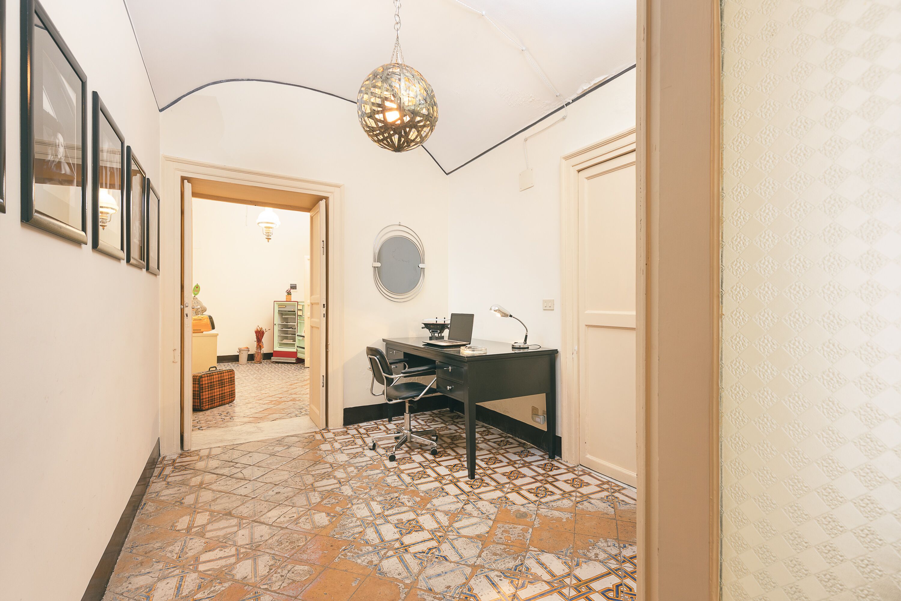 A hallway with a tiled floor, a desk with a chair, a hanging light fixture, and framed pictures on the wall.