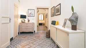 Un couloir étroit au sol recouvert de carreaux décoratifs, une commode avec une lampe, des tableaux encadrés aux murs et une sculpture originale posée sur un meuble.