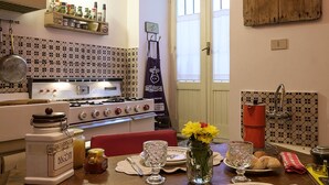 Une cuisine équipée d’une cuisinière, d’une planche à découper en bois sur laquelle se trouve du pain, et d’une table à manger dressée avec des assiettes et des couverts.