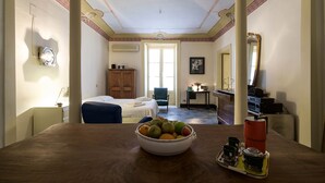 Une chambre d’hôtel avec une table à manger en bois, un panier de fruits, un plateau avec un thermos et un lit avec une chaise bleue.