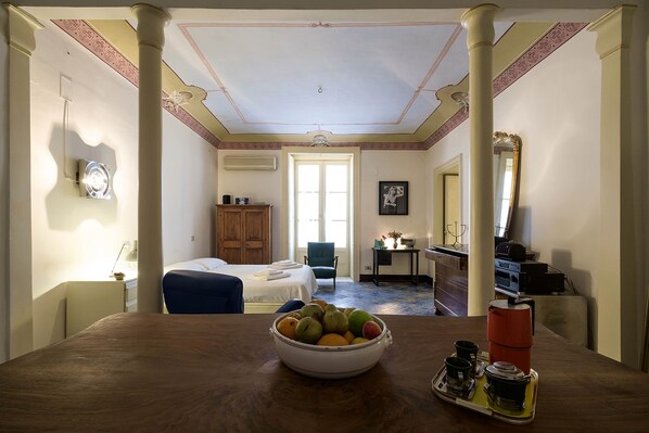 A hotel room with a wooden dining table, a bowl of fruit, a tray with a thermos, and a bed with a blue chair. - Casa Particular Luxury (Palermo)