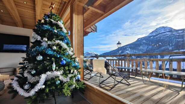 Una terraza de madera con un árbol de Navidad, un banco y vistas a montañas nevadas.