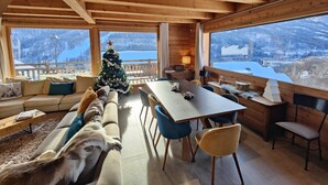 Interior acogedor de una cabaña con techo de madera, comedor, un árbol de Navidad y vista a un paisaje nevado.