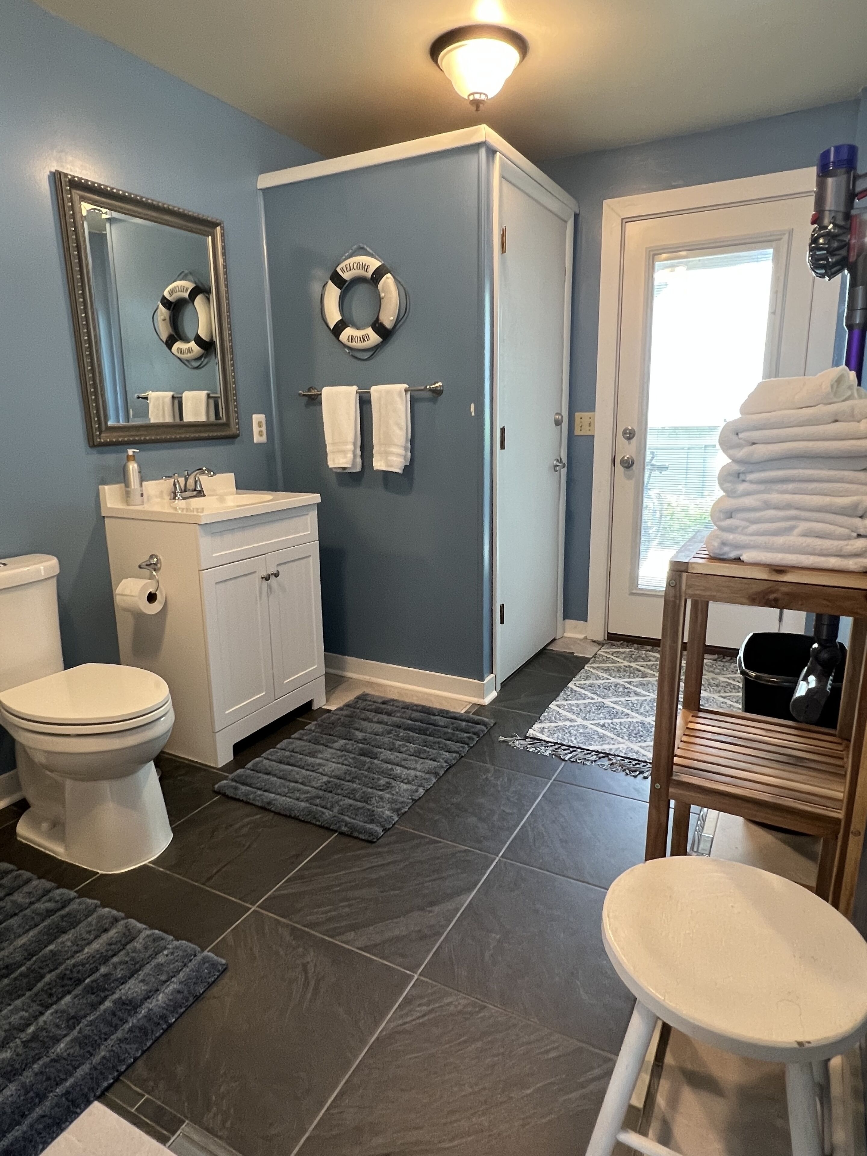A bathroom with a toilet, sink, and mirror. There are towels hanging on the wall and a stack of towels on a chair.