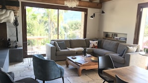 Un salon chaleureux avec un canapé gris, des fauteuils noirs, une table basse en bois et une vue sur la verdure à l’extérieur.