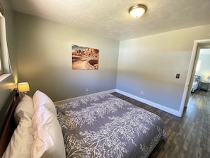 A bedroom with a bed, a nightstand, a lamp, a wall-mounted picture, and a door leading to another room. - Family Home in Kanab w/ private Hot Tub, Near Zion (Kanab)