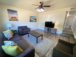 A living room with a sofa, armchair, coffee table, TV, and artwork on the walls. - Family Home in Kanab w/ private Hot Tub, Near Zion (Kanab)
