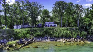 Um terreno à beira do lago com várias cabanas e uma casa grande, cercado por árvores densas e uma margem rochosa.