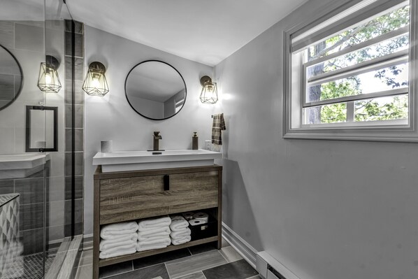 A modern bathroom with a large mirror, a round mirror, and three wall-mounted lights. There is a window with a view of trees and a shower area with glass doors.