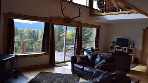 A spacious living room with a large window overlooking a deck and mountains. A leather sofa and a television are visible. - Elk Ridge (Packwood)