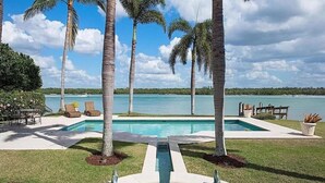 Zona de piscina con palmeras, vista al agua y una terraza con sillas de descanso.