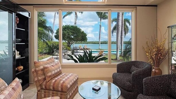 Una sala de estar con un amplio ventanal que ofrece vistas a un paisaje tropical y al mar. Hay dos sillones estampados y una mesa de centro de vidrio.