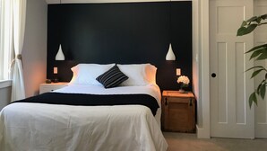 Ein Schlafzimmer mit einem großen Bett, zwei Nachttischlampen, einer gewebten Pendelleuchte, einem Nachttisch mit einer Vase und einer Tür mit einer Pflanze.