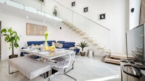 A modern living room with a glass dining table, blue sofa, and a staircase leading to an upper level.