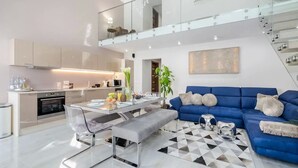 A modern kitchen with stainless steel appliances, a dining area with a glass table, and a blue sofa with decorative pillows.