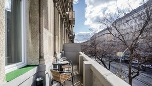 A balcony with a table and chairs, a window, and a view of a street with cars and trees.