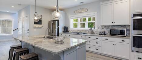 Una cocina moderna con armarios blancos, una isla grande con taburetes, electrodomésticos de acero inoxidable y un letrero que dice 'Beach House'.