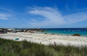 Une plage avec de gros rochers, une eau d’un bleu limpide et des bateaux.