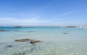 Une plage avec une eau claire et peu profonde, des rochers et une côte visible au loin.