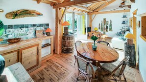 Een rustieke keuken met houten balken, een houten eettafel, rieten stoelen en uitzicht naar buiten.